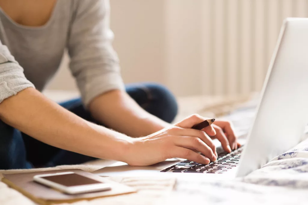unrecognizable-young-woman-sitting-on-bed-working-home-office