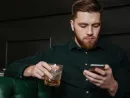 serious-man-sitting-and-drinking-whiskey-while-holding-cellphone