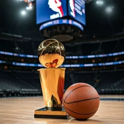 Indoor photo of photography of a basketball and next to it the larry o’brien nba championship trophy lying on a basketball court in an nba arena