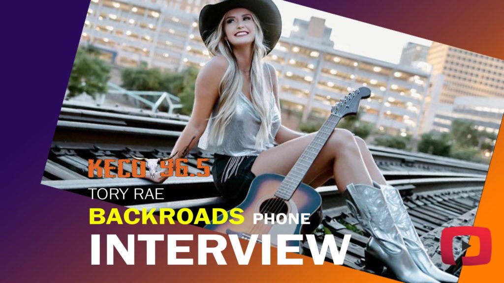 Tory Rae sitting on railroad tracks with her guitar, wearing cowgirl boots and a cowgirl hat.