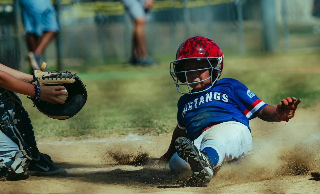 Child sliding into base during a baseball game wearing a Mustangs blue jersey and red helmet.