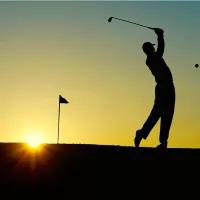 Golfer swinging club with ball in mid-air against sunset backdrop.