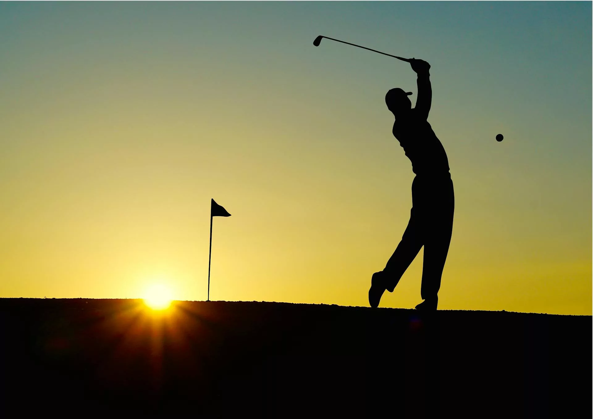 Golfer swinging club with ball in mid-air against sunset backdrop.