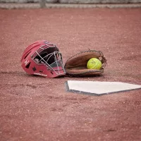 Crimson pitcher's mask, mitt with a softball, and home plate surrounded by dirt