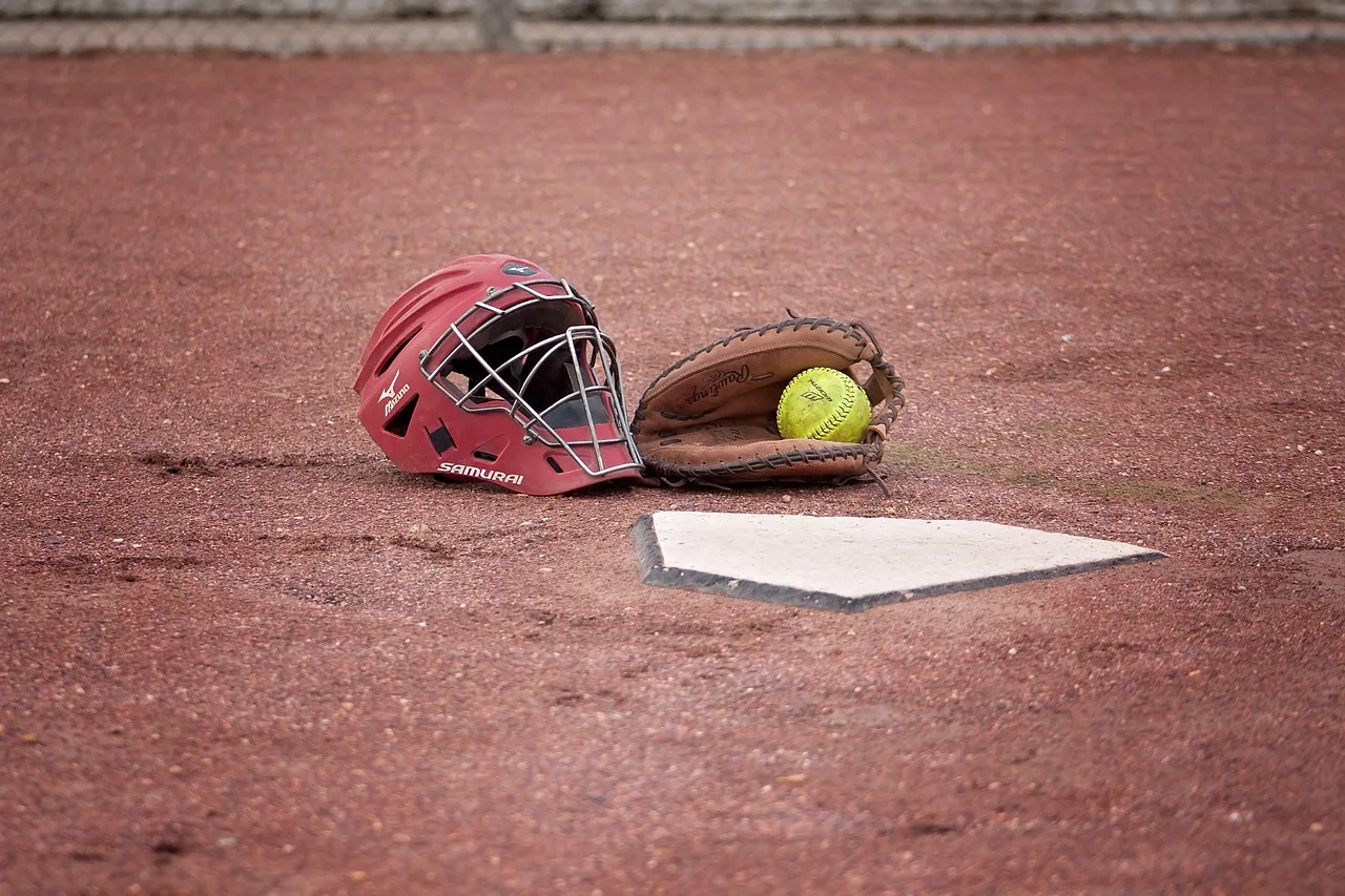 Crimson pitcher's mask, mitt with a softball, and home plate surrounded by dirt