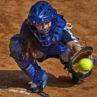 Teen female catcher in all blue catching a softball surrounded by dirt.