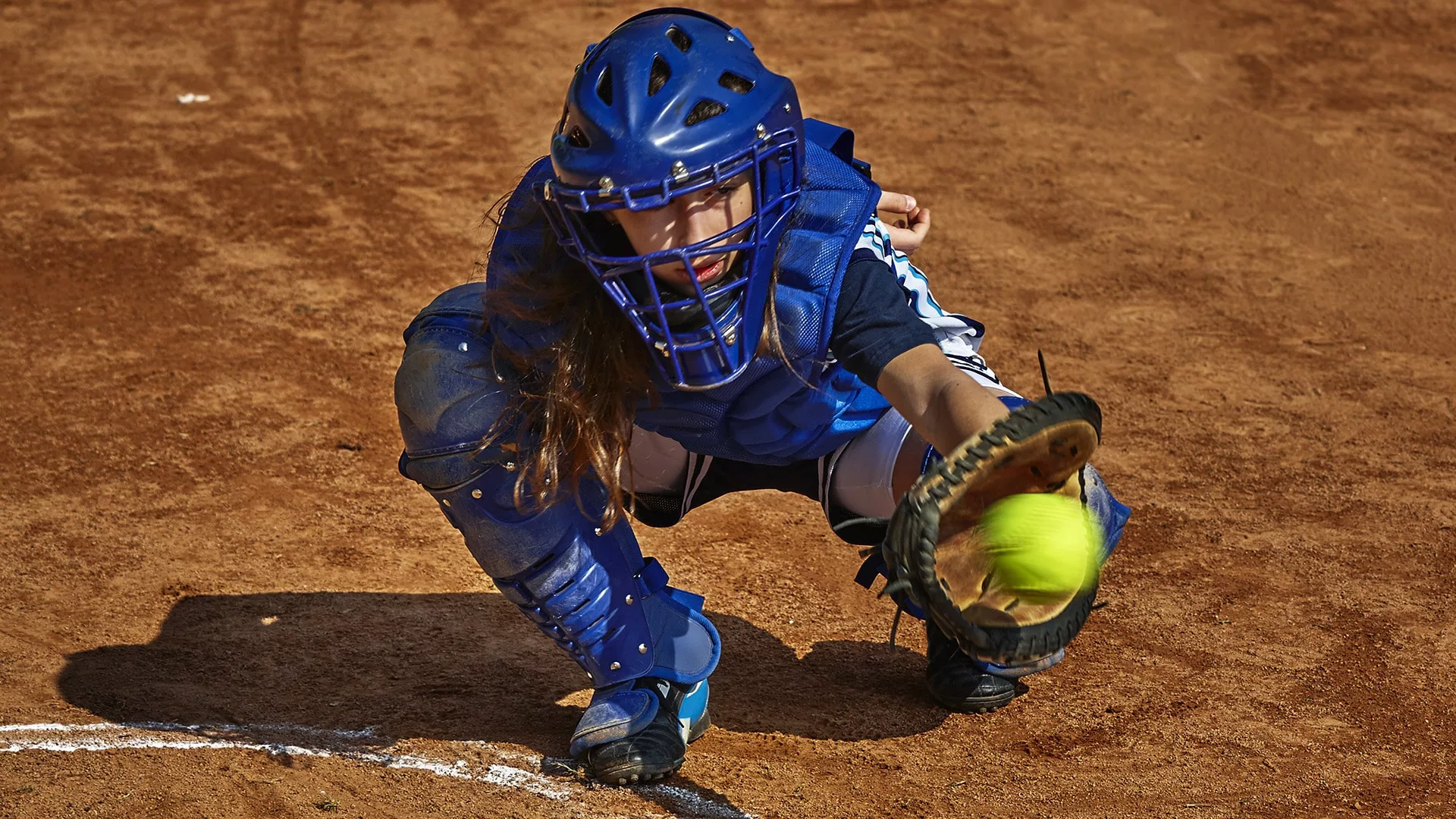 Teen female catcher in all blue catching a softball surrounded by dirt.