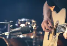 Male musician playing acoustic guitar behind microphone in recording studio.