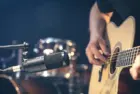 Male musician playing acoustic guitar behind microphone in recording studio.