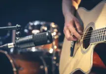Male musician playing acoustic guitar behind microphone in recording studio.
