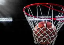 Looking up at an orange basketball falling through the rim and a white nylon net. With the arena lights in the background.