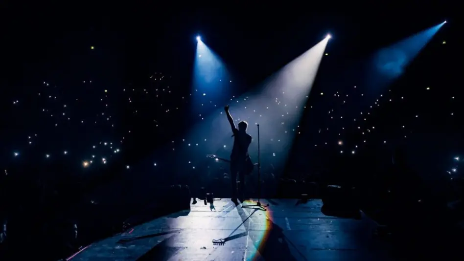 View from the stage stadium. Vocalist of a popular pop band on the background of the flashlights of phones during a concert. Fans switch on lights on smart phones on tribunes. Crowd waving cellphones