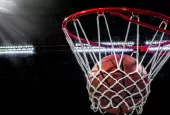 Looking up at an orange basketball falling through the rim and a white nylon net. With the arena lights in the background.