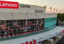 Aerial 4K drone shot captures a high-angle twilight view of the Lenovo Center arena^ home of the NHL's Carolina Hurricanes. The low golden hour sun rakes across the modern facade^ highlighting the reflective teal glass panels of the Carolina Hurricanes home venue before they face the Montreal Canadiens. The shot documents the wide^ geometric infrastructure layout of the empty parking lots^ main entrance gates^ and surrounding roadway logistics under a clean sunset gradient sky. Raleigh^ North Carolina^ USA - May 19^ 2026
