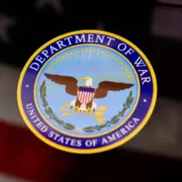 The United States Department of War seal is displayed on a smartphone screen. In the background^ an out-of-focus American flag is visible. This conceptual image illustrates themes of US military history^ national defense^ government institutions^ and historical conflicts.
