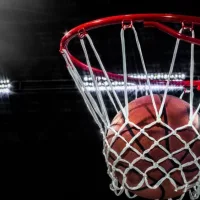 Looking up at an orange basketball falling through the rim and a white nylon net. With the arena lights in the background.