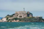 Alcatraz Island in San Francisco Bay^ California^ USA^ viewed from the water. Shows rugged terrain^ prison building^ lighthouse^ and choppy water under clear sky.
