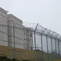 Looking up at the outside walls of a modern prison surrounded by a fence and barbed wire.