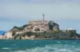 Alcatraz Island in San Francisco Bay^ California^ USA^ viewed from the water. Shows rugged terrain^ prison building^ lighthouse^ and choppy water under clear sky.