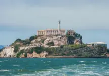 Alcatraz Island in San Francisco Bay^ California^ USA^ viewed from the water. Shows rugged terrain^ prison building^ lighthouse^ and choppy water under clear sky.