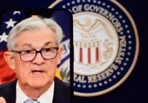 Federal Reserve (Fed) Chair Jerome Powell is visible on smartphone screen. In the background^ an inflation rate chart is displayed.