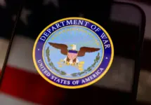 The United States Department of War seal is displayed on a smartphone screen. In the background^ an out-of-focus American flag is visible. This conceptual image illustrates themes of US military history^ national defense^ government institutions^ and historical conflicts.