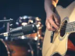 Male musician playing acoustic guitar behind microphone in recording studio.