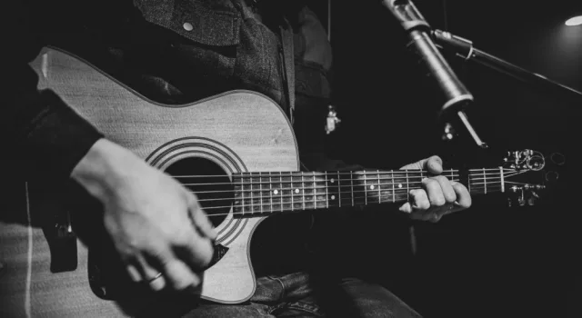The guitarist plays an acoustic guitar with a capo in front of a microphone. The concept of music recording^ rehearsal or live performance.