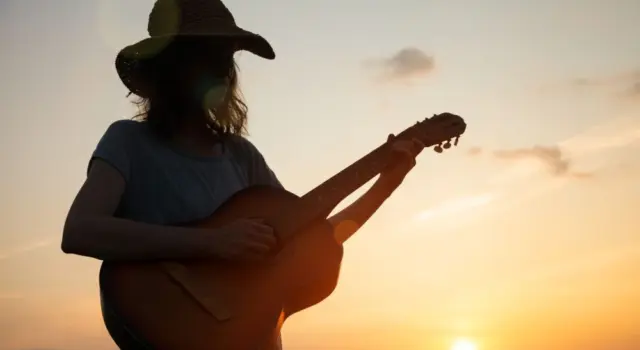silhouette of young free woman in straw hat playing country music on a guitar at sunset^ copy space