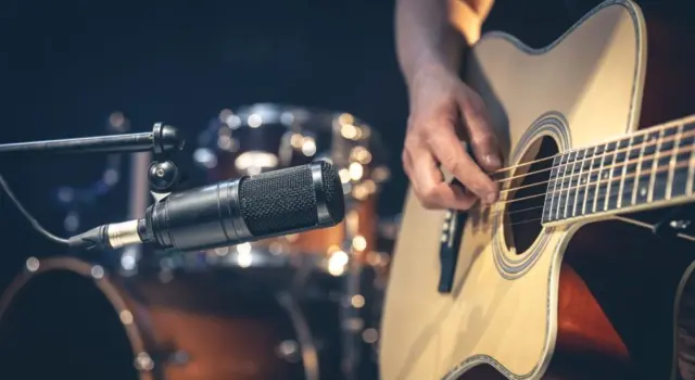Male musician playing acoustic guitar behind microphone in recording studio.