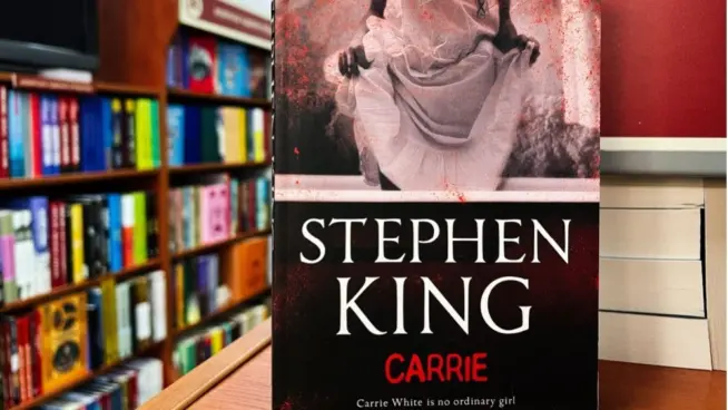 Paperback cover of Carrie by Stephen King shown on a bookshop shelf background