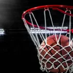 Looking up at an orange basketball falling through the rim and a white nylon net. With the arena lights in the background.