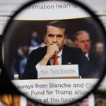 Close up through magnifying lens capturing legal professional Todd Blanche with nameplate at desk during hearing. Law and courtroom justice journalism concept.