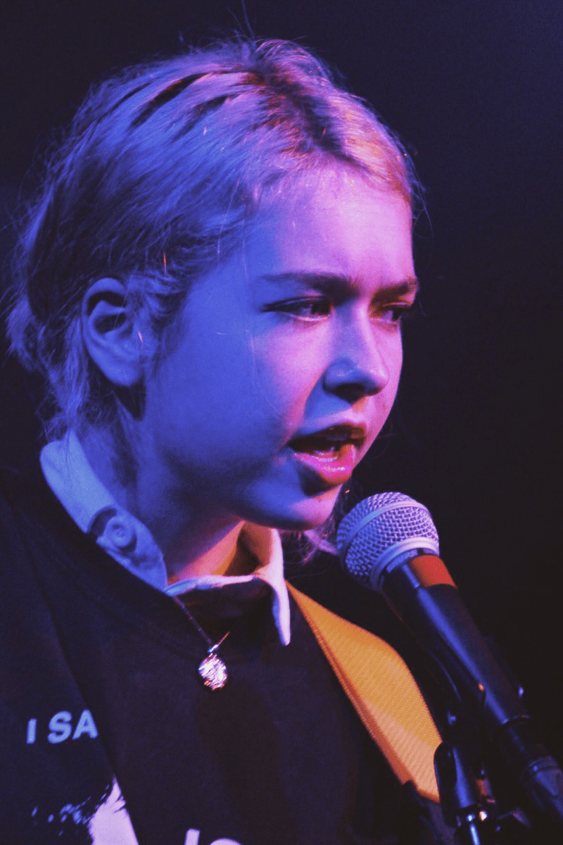 A close up photo of the lead singer of Snail Mail, Lindsey Jordan. She is bathed in a purple light and sings softly into a microphone