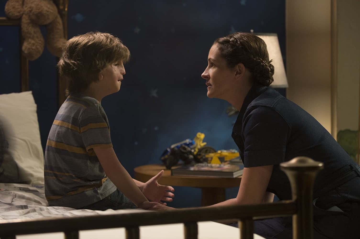 Actor Jacob Tremblay as August Pullman is sitting on his bed while Julia Roberts, who plays his mother Isabel Pullman, kneels down to speak to him 