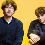 Stephen and Erik Paulson sit in front of a yellow background, looking at the camera