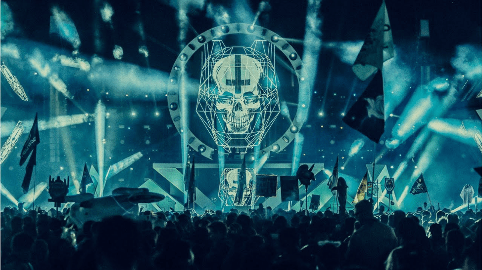 Photo from within the crowd at a concert where BTSM is performing. Blue lights fill the space with white spotlights shining into the crowd and behind the trio that is performing on stage there is a geometric large skull.