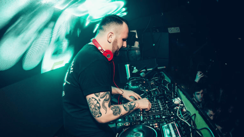 Photo of Zomboy mixing beats in a black T shirt with red beats around his neck. He has black tattoos on his right arm and is lit by green- blue lights behind him, and to his right is the audience.