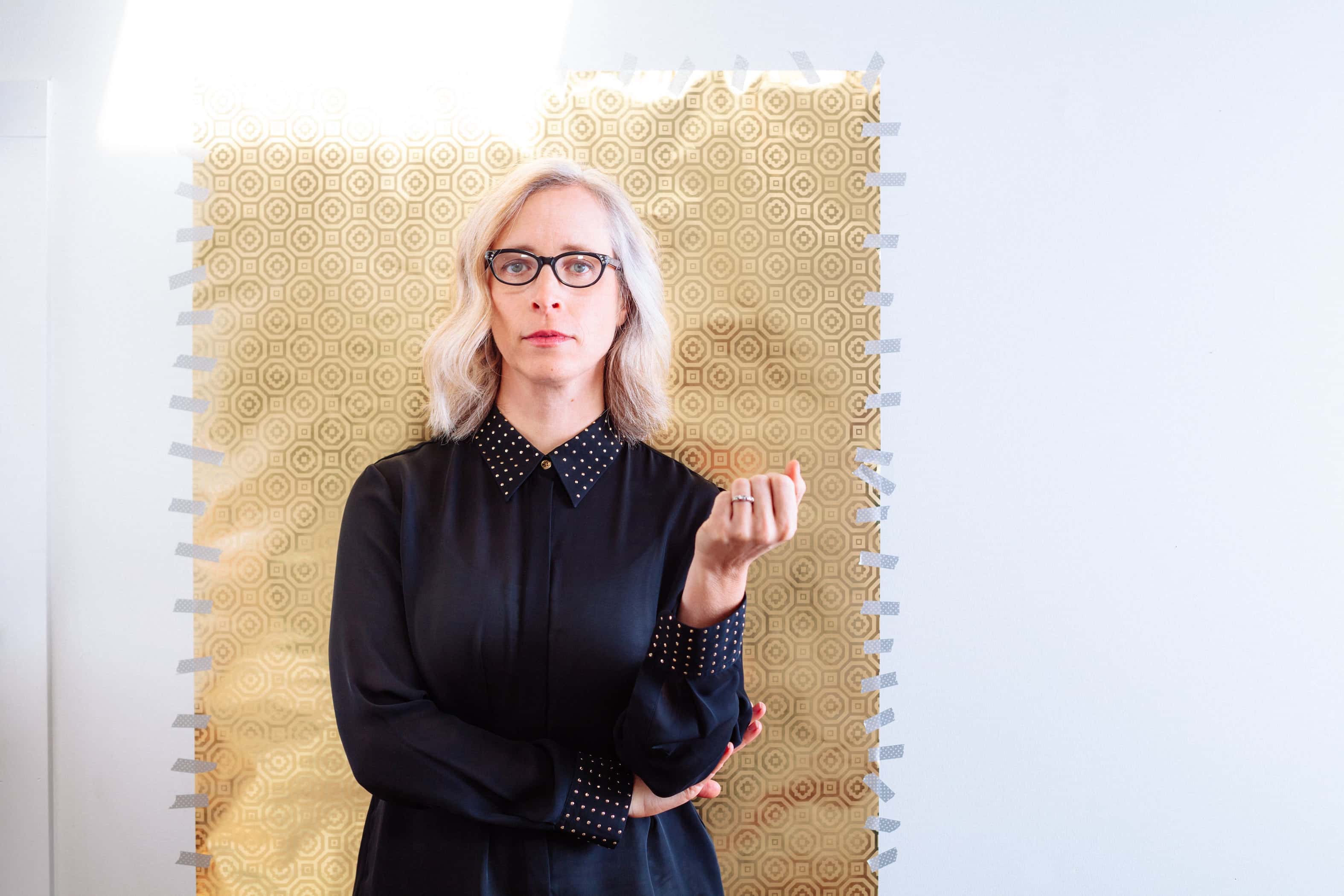 Laura Veirs (blonde hair and wearing a black button-up shirt) stands in front of a white and gold background.