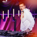 Matoma mixing beats on a mixing table, wearing a white hoodie, illuminated by pink and orange lights.