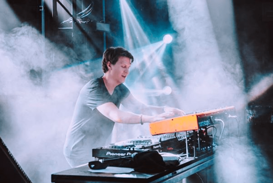Matoma using a mixing board and keyboard to create music. He is surrounded by white lights and dry ice that make fog.