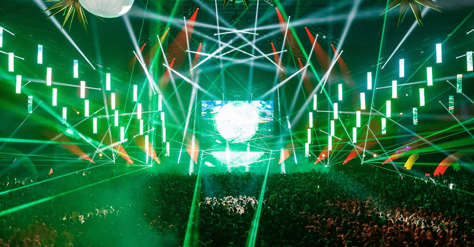 A large stage in front of a crowd, green light beams illuminate the concert goers.