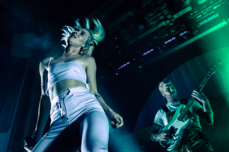 Georgia, wearing a white crop top and white jeans, dances on stage as her brother Caleb plays guitar behind her. 