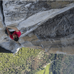 Alex Honnold is high above Yosemite National Park in the middle of his ascent of El Capitan. The camera is looking down at him with as he is crack climbing on the granite wall. In the distance below him is a forest of deep green trees