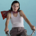 King Princess sits atop a bike in front of a blue backdrop and wearing a red scarf