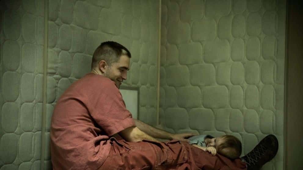 The main character Monte (played by Robert Pattinson, sits in a futuristic prison cell with a small child in his lap. They are both smiling at one another