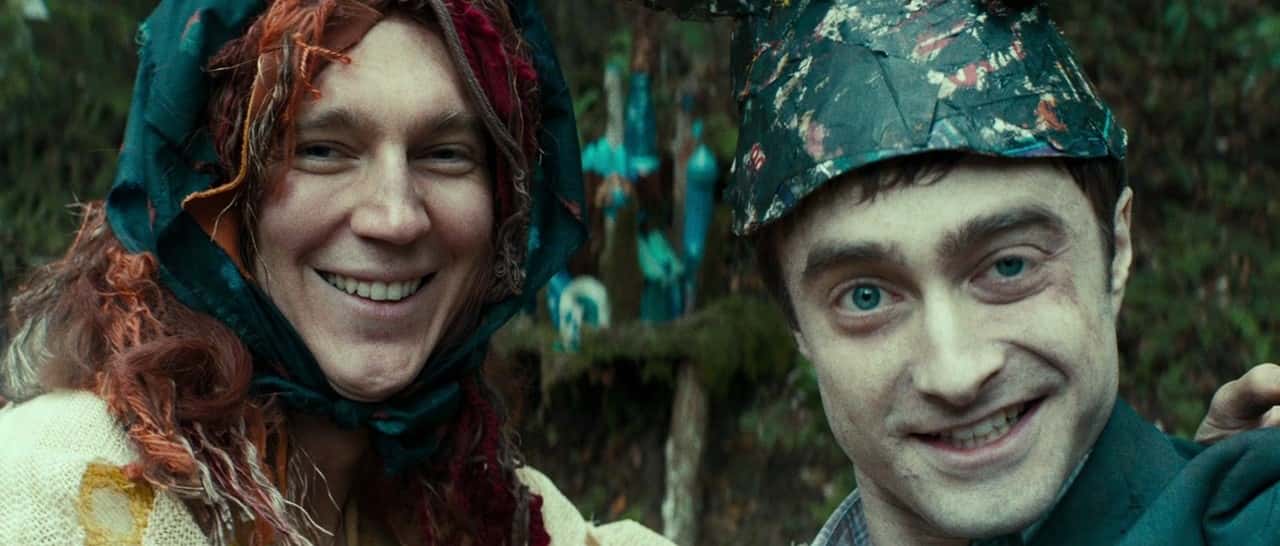 Paul Dano is dressed in a wig and knitted sweater with his arm around Daniel Radcliffe who is wearing a homemade hat. They are posing for a selfie in the forest.