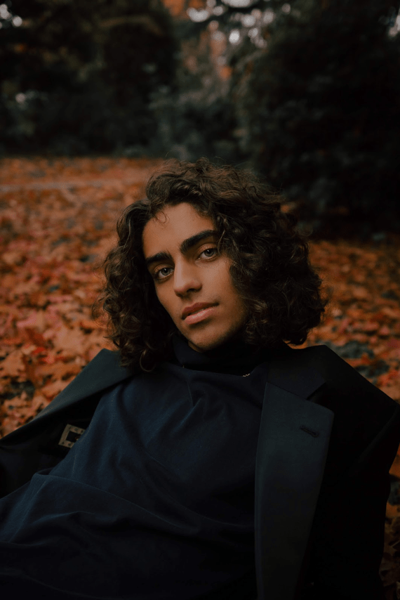 A headshot of a male model in all black sits in orange fall leaves