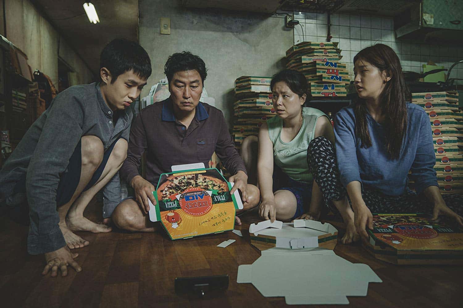 The Kim family sitting on their kitchen floor with piles of freshly folded pizza boxes behind them and more in their hands. They all look exhausted and disheveled.