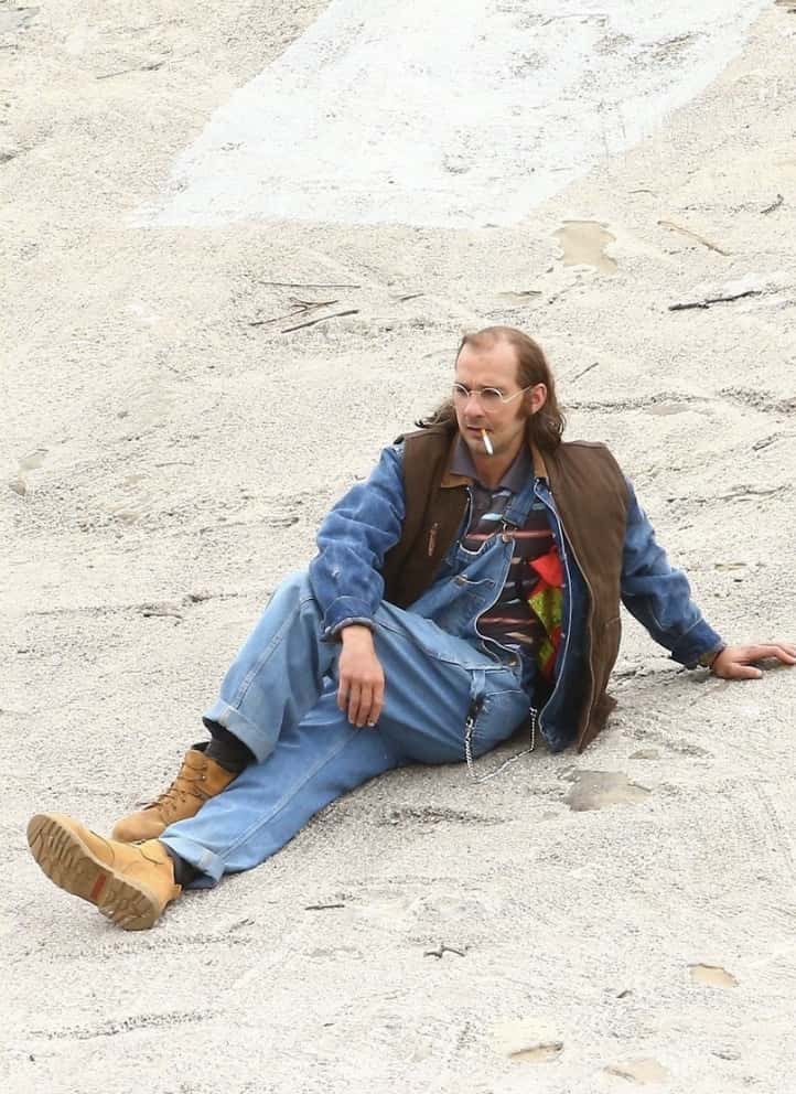 Shia LeBeouf pictured as his character James Lort. He is sitting on the ground, cigarette dangling from his mouth with loosely fitted clothes and a receding hairline.